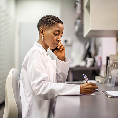 Physician writing at a desk while holding a phone to her ear.